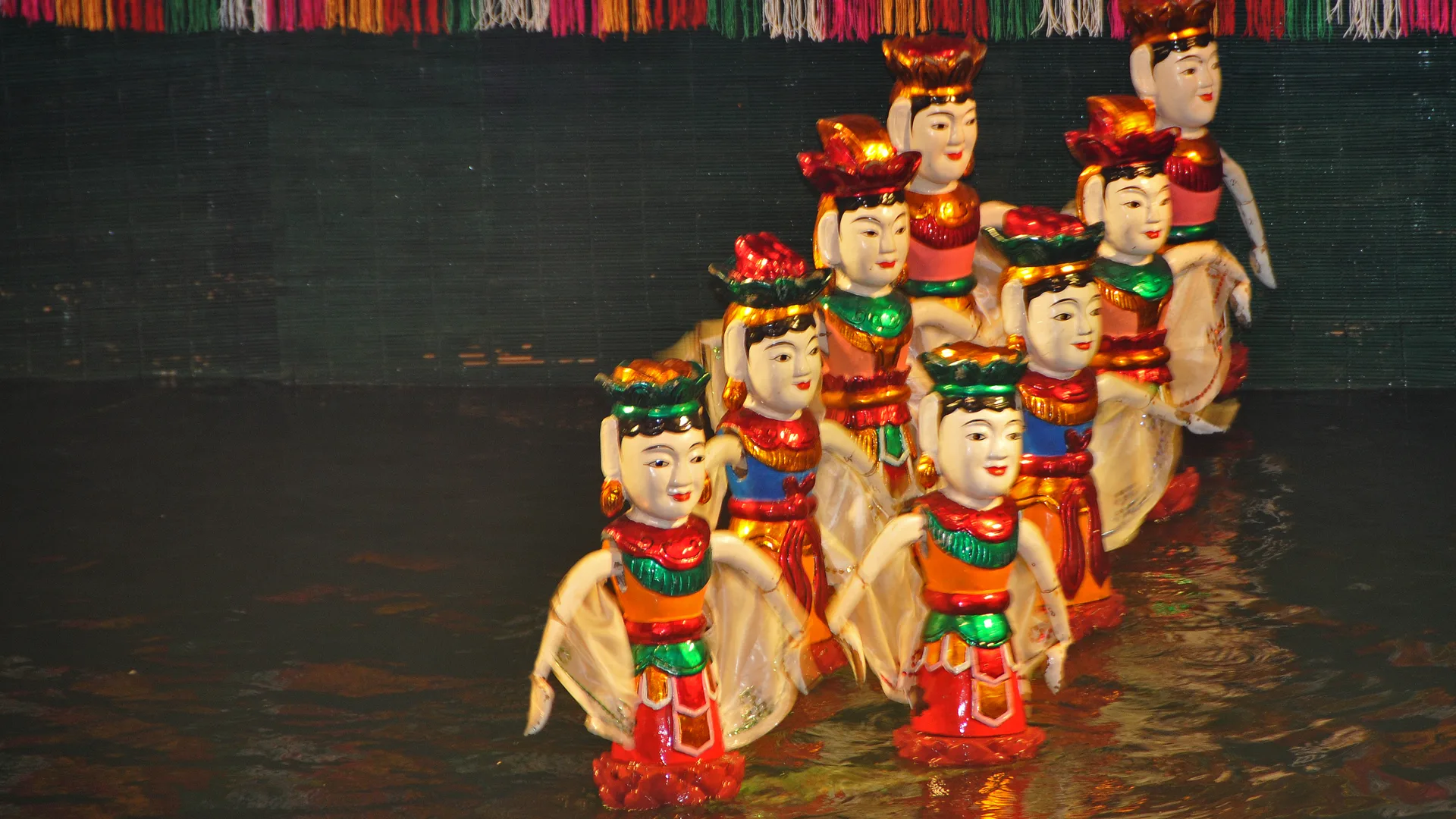 A group of lacquered wooden water puppets with painted faces, lotus-shaped headdresses and orange, red and green costumes standing in dark water before a green bamboo screen