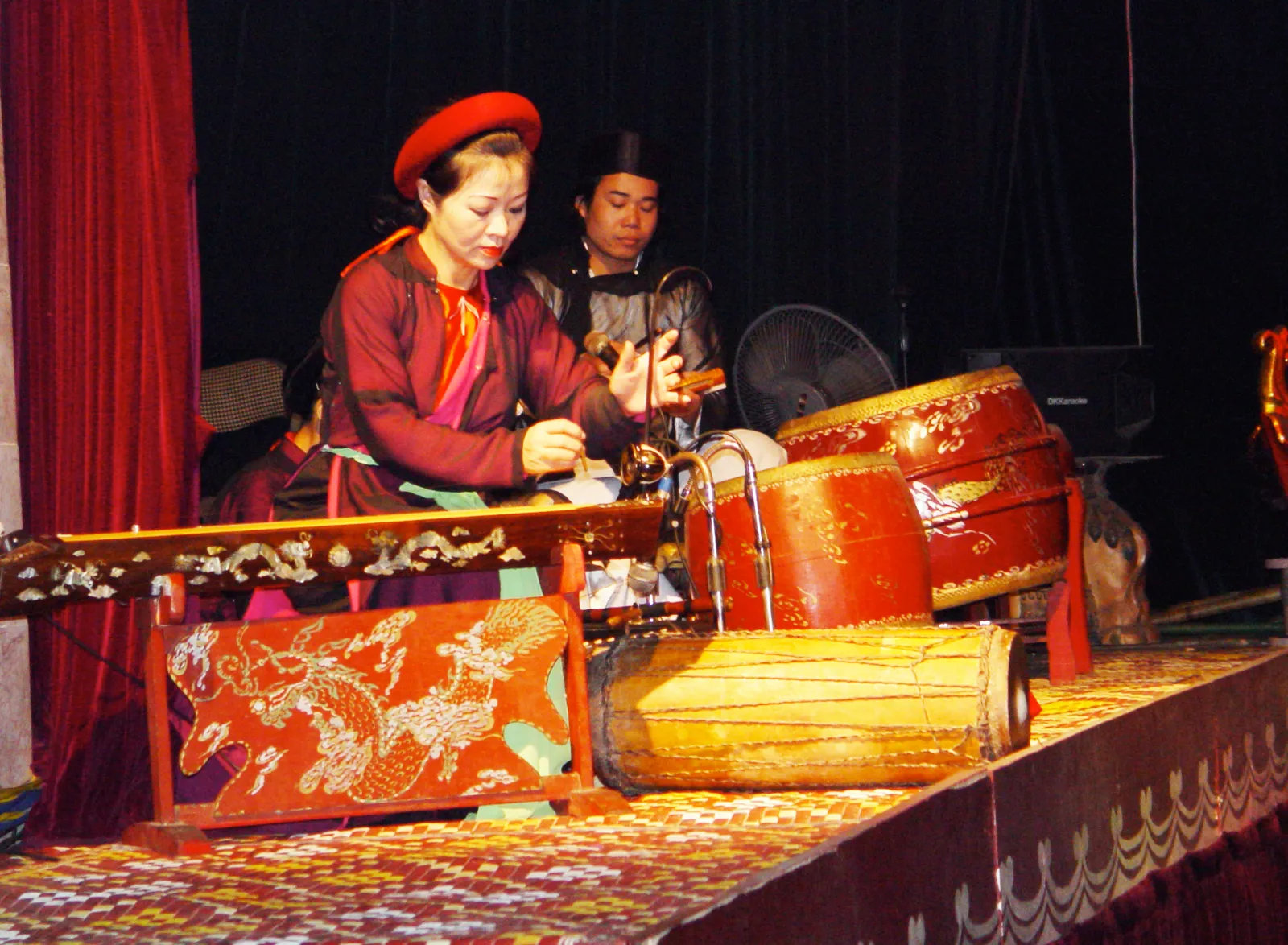 A musician in a red hat and traditional dress playing beside drums and a decorated instrument on the side of a water-puppet stage; a show in Hanoi