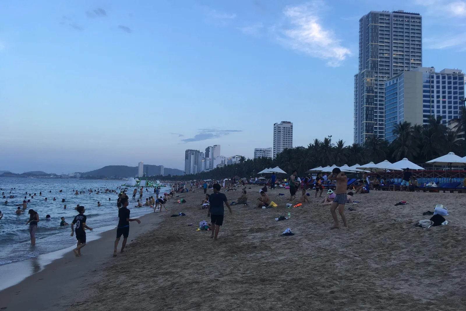 People walking and paddling on a wide sand beach at dusk, with beach umbrellas, palms and tall hotel towers behind, in Nha Trang