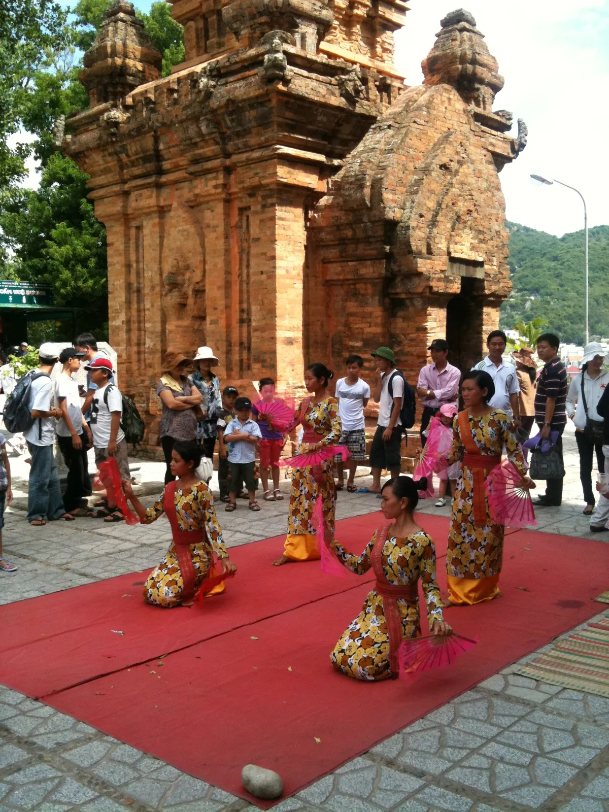 Dancers in orange and gold costumes kneeling on a red mat in front of a carved brick Cham tower, with onlookers around them
