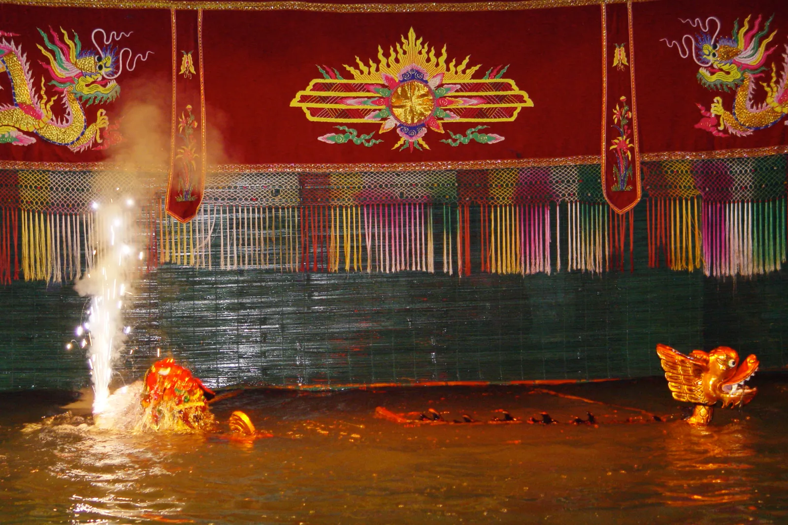 A water-puppet stage with a red embroidered curtain, a green bamboo screen and a fountain of sparks beside a gold puppet dragon head on the water; a show in Hanoi